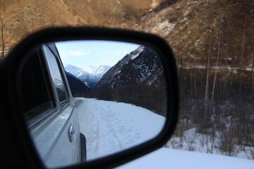 Reflection of snow-capped mountains and a snow-covered road in the rear-view mirror of a car,...