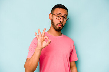 Young hispanic man isolated on blue background winks an eye and holds an okay gesture with hand.