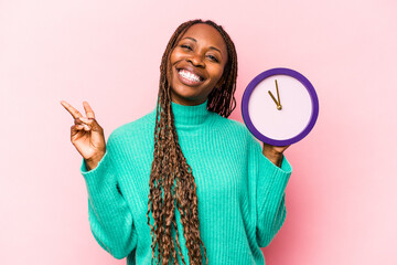 Young African American woman holding a clock isolated on pink background joyful and carefree showing a peace symbol with fingers.