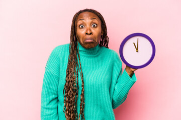 Young African American woman holding a clock isolated on pink background shrugs shoulders and open...