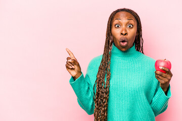 Young African American woman holding an apple isolated on pink background pointing to the side