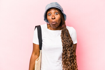 Young African American woman going the beach isolated on pink background shrugs shoulders and open...
