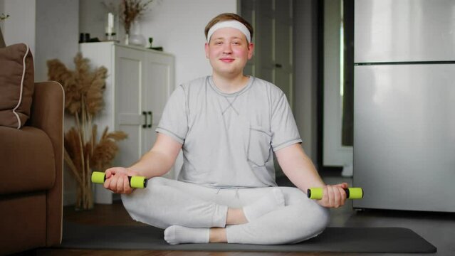 A chubby guy in sportswear performs exercises with dumbbells while sitting on a mat. Sport