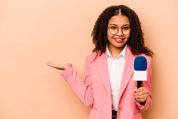 Young African American TV presenter woman isolated on beige background showing a copy space on a palm and holding another hand on waist.