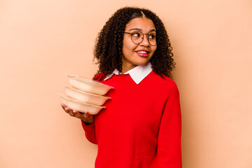 Young African American woman holding tupperware isolated on beige background looks aside smiling,...