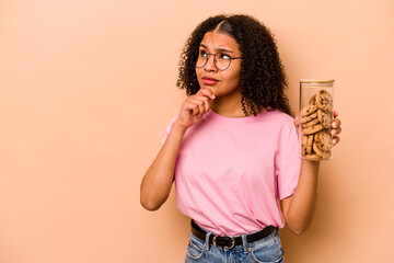 Young African american woman holding a cookies jar isolated on beige background looking sideways with doubtful and skeptical expression.