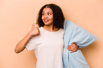 Young African American woman holding a pillow isolated on beige background points with thumb finger away, laughing and carefree.