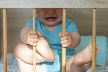 cute little baby boy is crying in the crib, holding the wooden bars with hands and pushing with...