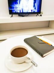 Coffee mug on white round table, there is a notebook and pen on the right side. Next is a table with a monitor. clean cream color background.