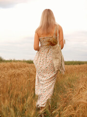 Woman walks through a wheat field in a sundress with a linen bag. Wheat spikelets stick out of the bag. Copy space. Walk to the yellow wheat field, beautiful rural landscape. Rear view. Copy space
