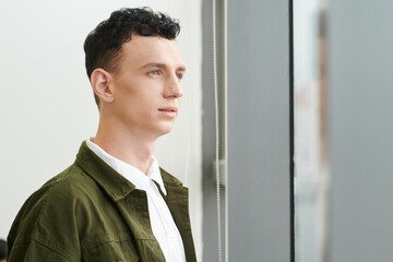 Portrait of pensive ambitious young man with curly hair looking outside through window