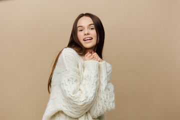 a cheerful woman is standing sideways on a light brown background in a white sweater, smiling sweetly with her hands touching each other to her face