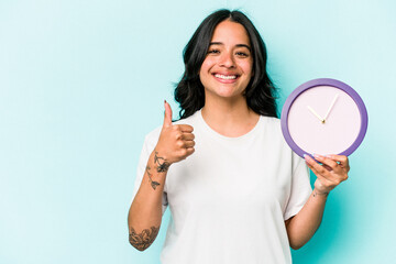 Young hispanic woman holding a clock isolated on blue background smiling and raising thumb up