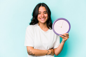 Young hispanic woman holding a clock isolated on blue background laughing and having fun.