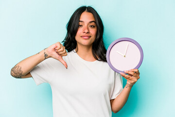 Young hispanic woman holding a clock isolated on blue background showing a dislike gesture, thumbs down. Disagreement concept.