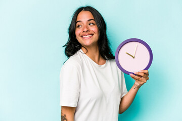 Young hispanic woman holding a clock isolated on blue background looks aside smiling, cheerful and pleasant.