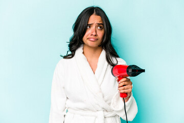 Young hispanic woman holding dryer isolated on blue background confused, feels doubtful and unsure.