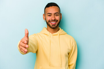 Young hispanic man isolated on blue background stretching hand at camera in greeting gesture.