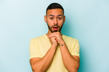 Young hispanic man isolated on blue background praying for luck, amazed and opening mouth looking to front.
