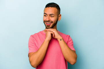 Young hispanic man isolated on blue background praying for luck, amazed and opening mouth looking to front.