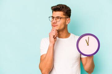 Young caucasian man holding a clock isolated on blue background looking sideways with doubtful and...