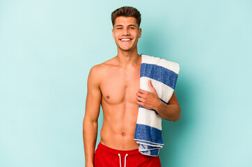 Young caucasian man holding beach towel isolated on blue background happy, smiling and cheerful.