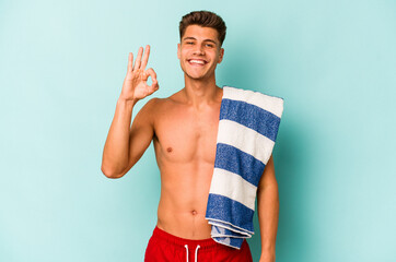 Young caucasian man holding beach towel isolated on blue background cheerful and confident showing...
