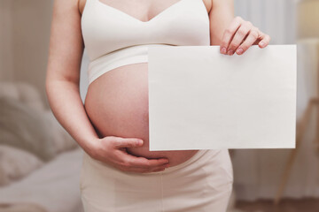 Pregnant woman with a sheet of A4 paper in her hands, home living room