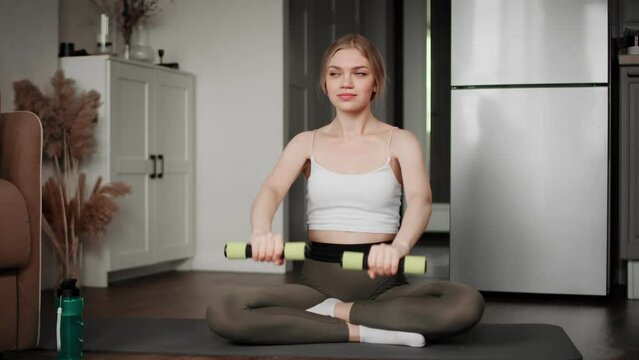 A girl in sportswear performs exercises with dumbbells, sitting on a mat. Sport