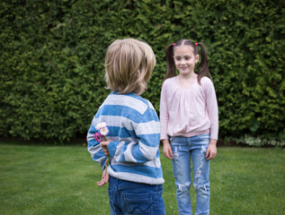 Fototapeta premium boy hid daisy flowers behind his back, surprise for his little friend. A defocused girl is waiting. The concept of friendship and child love. Unexpected, pleasant childish sign of attention