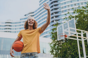 Bottom view young man 20s yellow t-shirt bandana waving hand say hello playing basketball hold ball on playground in free time rest relax in city outdoors on open air. Urban lifestyle leisure concept. © ViDi Studio