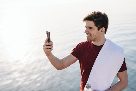 Young strong sporty athletic fit sportsman man in sports clothes do selfie shot on mobile cell phone warm up training at sunrise sun dawn over sea beach outdoor on pier seaside in summer day morning.
