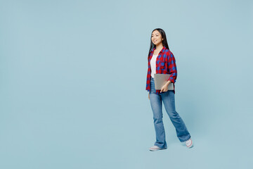 Full body young smiling fun woman of Asian ethnicity wear checkered shirt hold closed laptop pc computer walking going isolated on plain pastel light blue color background. People lifestyle concept.