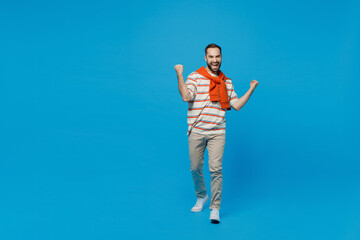 Full body young excited overjoyed man 20s wear orange striped t-shirt doing winner gesture celebrate clench fists say yes isolated on plain blue background studio portrait. People lifestyle concept.