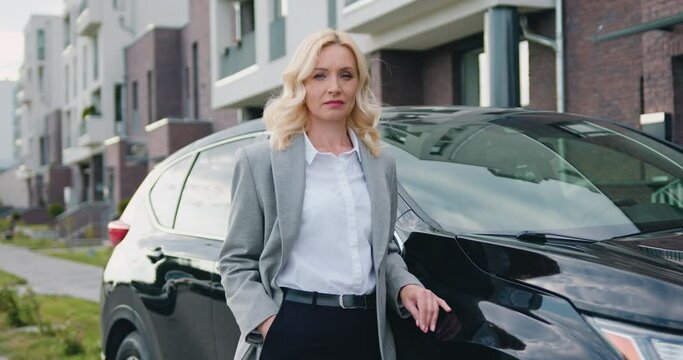 Portrait of successful young caucasian businesswoman in official clothes outside when standing beside black car on parking near modern building. Outdoors.