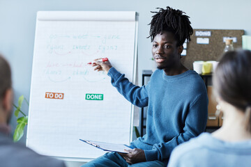 Portrait of young black man teaching English class in office to group of people and pointing at whiteboard with grammar rules - obrazy, fototapety, plakaty