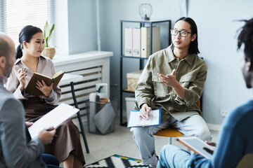Portrait of young Asian man asking audience during English lesson with diverse group of people sitting in circle in office setting