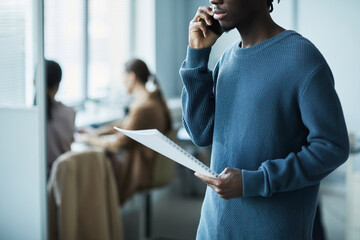 Cropped shot in blue of young African American man speaking on phone in office setting and holding...