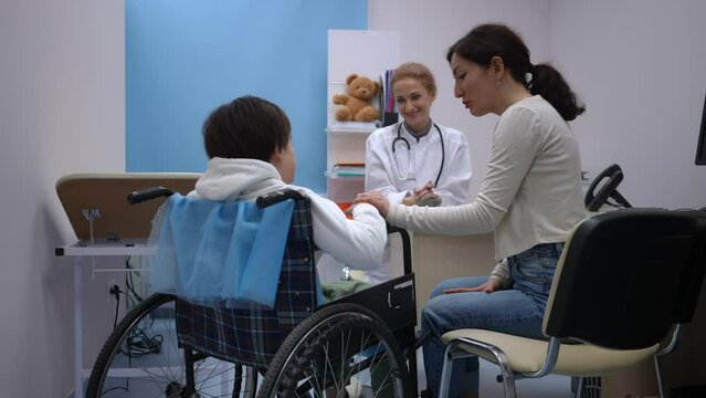 Back view smiling Caucasian mother holding hand of son in wheelchair talking as smiling doctor sitting at background. Positive woman endorsing supporting ill boy consulting professional specialist