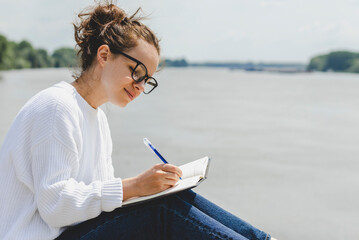 Woman wearing white sweater and glasses writing her ideas in her diary.