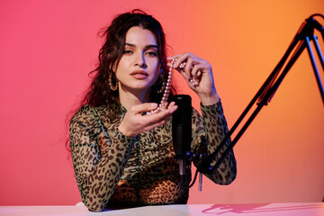 Modern young female blogger sitting in studio in pink and yellow neon light recording sounds for ASMR vlog using bead necklace