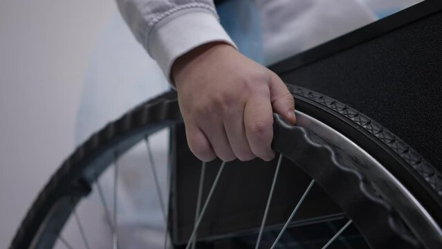 Close-up wheelchair wheel with unrecognizable Caucasian boy hand indoors. Little ill patient sitting on mobility aid device. Health care and disability concept