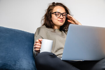 Young brunette woman using laptop and drinking coffee while sitting on sofa at home.