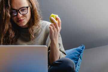 Smiling girl with glasses eating apple and looking at laptop.