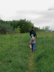 Family walking with dogs on green grass rural landscape. Countryside cottagecore style. Camping activity spring forest. Candid authentic people father and daughter with pets from behind hiking outdoor