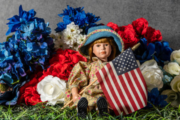 doll sitting on grass surrounded by red white and blue flowers holding American flag