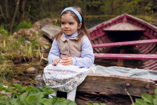 Little 3-eyar-old girl sitting in old wooden pink boat in village. Little baby girl of 3-years-old in retro vintage dress and vest smiling
