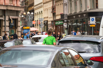 The upper body of a cyclist in a green T-shirt on a city road surrounded by cars and passers-by on the sidewalk against the background of the walls of houses in blur