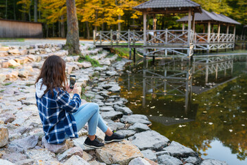 Woman traveler in a plaid shirt makes a photo of the lake with a mobile phone. Beautiful autumn forest.