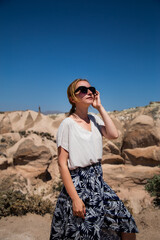 Fototapeta premium Young happy woman traveler enjoying a vacation in desert Cappadocia Turkey. Tourist girl in sunglasses and dress outdoor portrait. Sunny female portrait in the desert.
